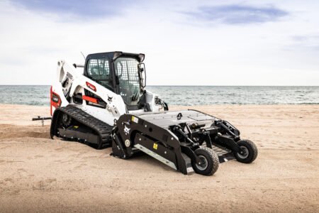 White Bobcat skid-steer loader on a sandy beach with the sea and blue sky in the background for context and scale.