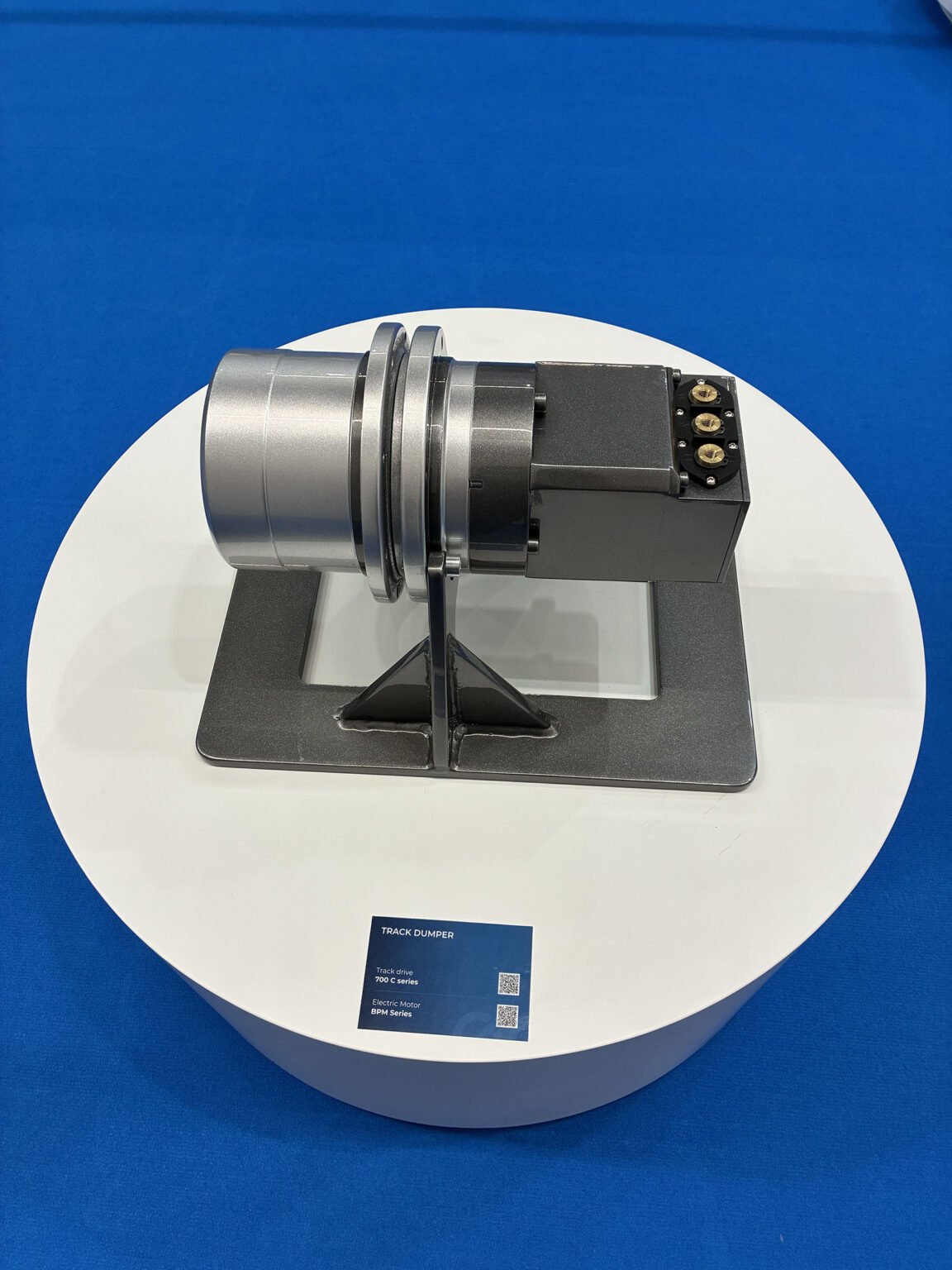 Bonfiglioli-IVT-Expo-3jpg - Machines Wiki Track dumper demonstration unit: a silver cylindrical housing and black control box mounted on a metal stand, displayed on a white circular pedestal against a blue background.