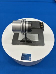 Track dumper demonstration unit: a silver cylindrical housing and black control box mounted on a metal stand, displayed on a white circular pedestal against a blue background.