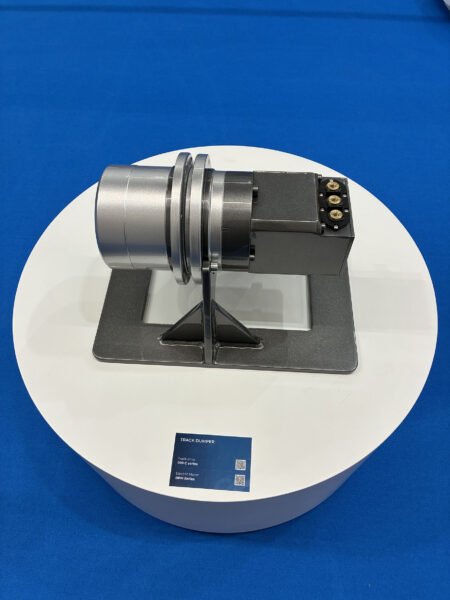 Bonfiglioli-IVT-Expo-3jpg - Machines Wiki Track dumper demonstration unit: a silver cylindrical housing and black control box mounted on a metal stand, displayed on a white circular pedestal against a blue background.