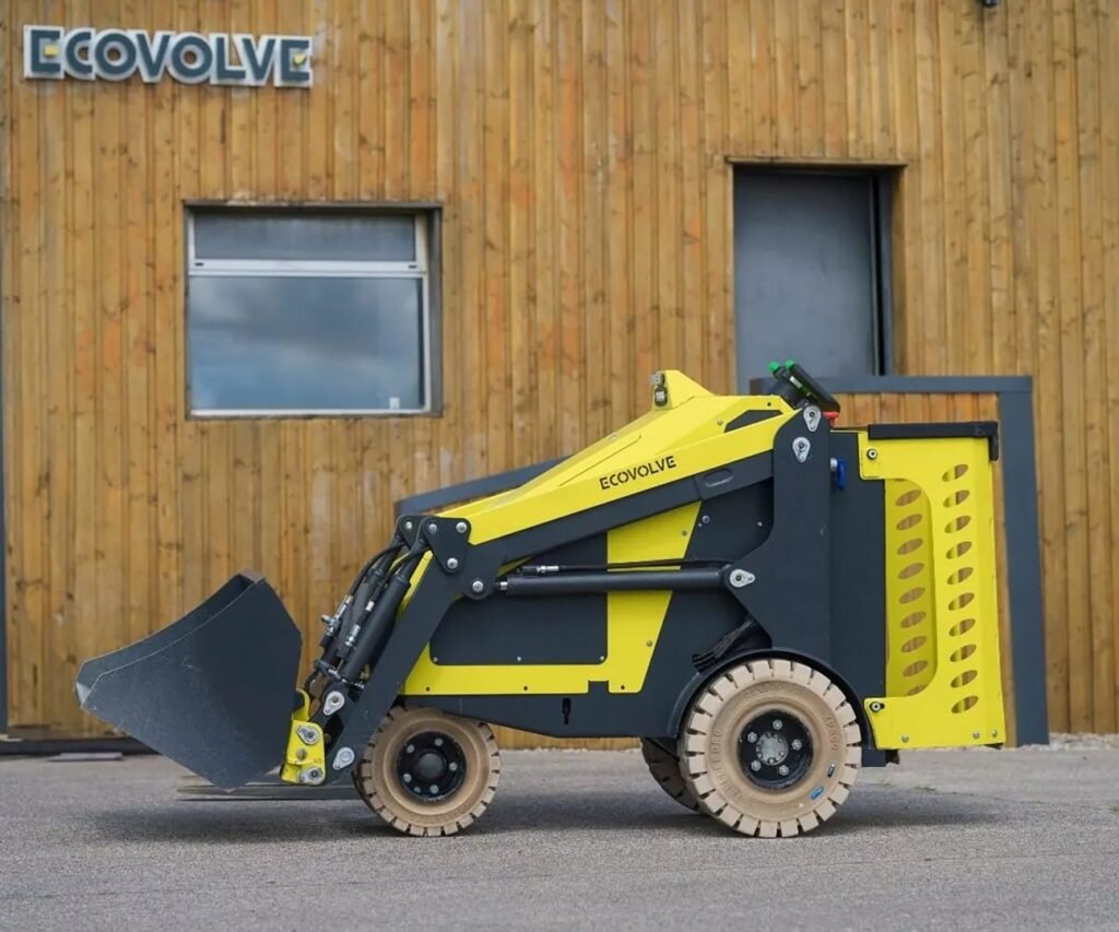 Yellow compact skid-steer loader with a front bucket parked on a paved surface in front of a wood-clad building with ECOVOLVE signage
