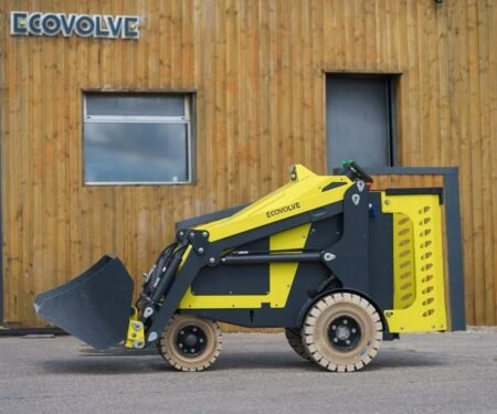Yellow compact skid-steer loader with a front bucket parked on a paved surface in front of a wood-clad building with ECOVOLVE signage