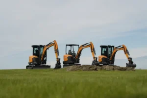 Three orange compact excavators lined up on a grassy field with a rocky pile and a clear blue sky above.