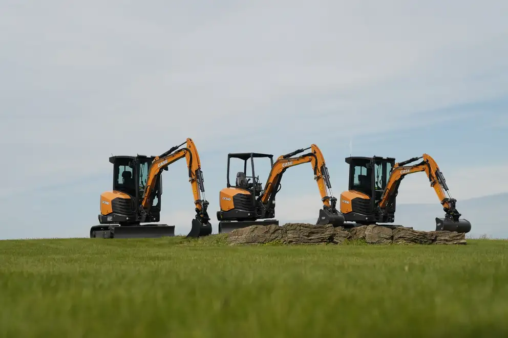 CASE Unveils Three New D Series Mini Excavators Three orange compact excavators lined up on a grassy field with a rocky pile and a clear blue sky above.