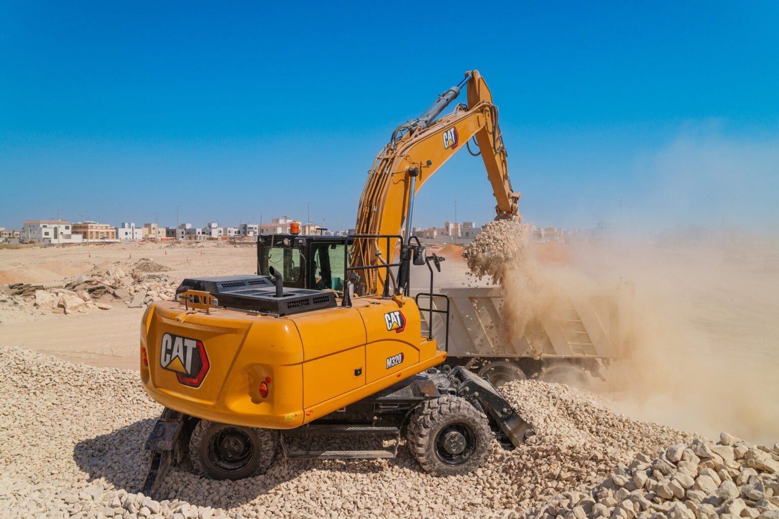 Yellow CAT excavator at a rocky construction site under a blue sky, with houses in the background.