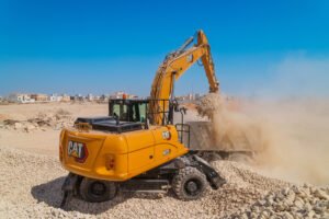 Yellow CAT excavator at a rocky construction site under a blue sky, with houses in the background.