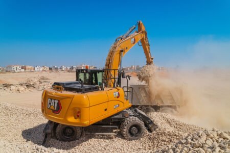 Cat-M320-WHEX-CM20251117-822d0-562ac-scaledjpg - Machines Wiki Yellow CAT excavator at a rocky construction site under a blue sky, with houses in the background.
