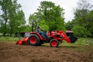 Man operating a red Kioti tractor with a front loader/backhoe attachment on a dirt field, trees in the background