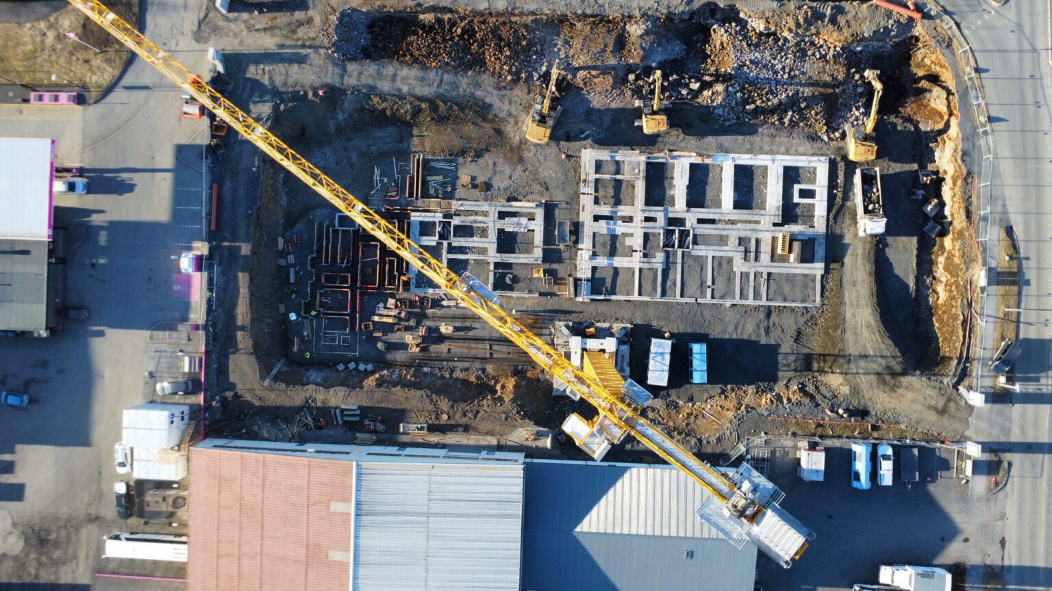 First-Potain-MDT-489-in-Iceland-drives-new-construction-thinking-on-Reykjavik-project-02-scaledjpg - Machines Wiki Aerial view of a construction site with a large yellow crane over a partially built concrete foundation and surrounding roads at the edges.