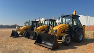 Gehl_dealer_South_Gear_machines-scaledjpg - Machines Wiki Row of yellow backhoe loaders with large front buckets lined up in a dirt lot under a clear blue sky near a white industrial building, GEHL machines visible.