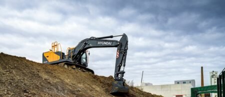 HX230-Web-BannerLowjpg - Machines Wiki Hyundai hydraulic excavator on a dirt mound at a construction site, with cloudy sky and industrial buildings in the background.