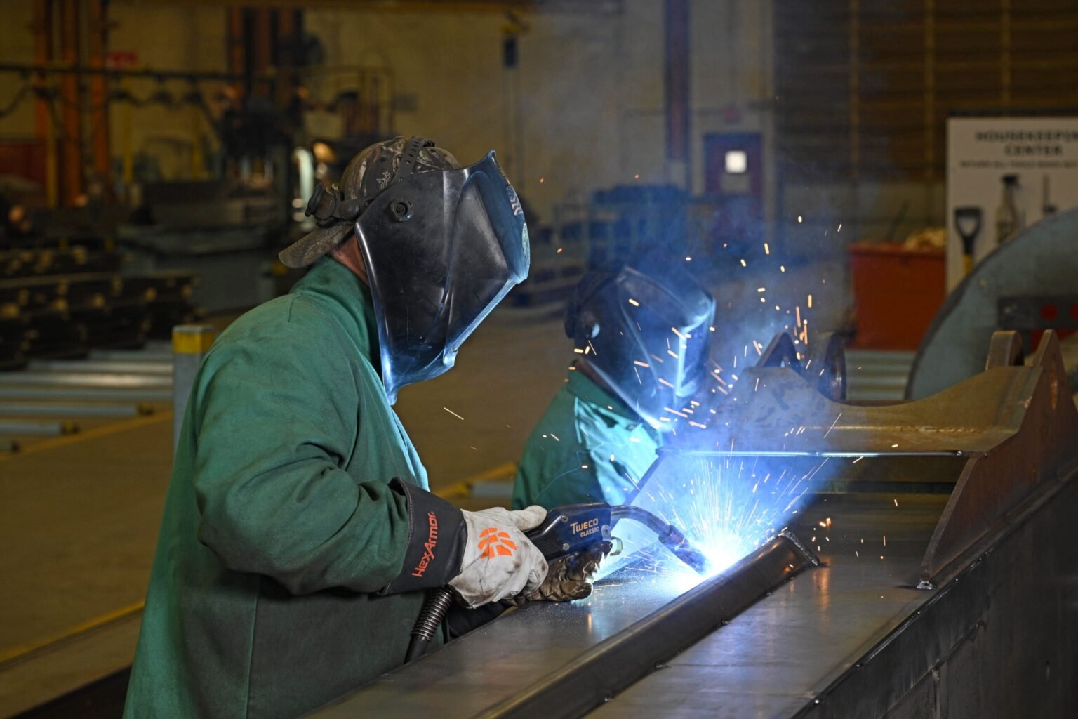 Manitowoc-expands-US-aftermarket-support-with-enhanced-Rapid-Response-Shop-01-scaledjpg - Machines Wiki Welder in a green protective jacket and helmet welding metal, sparks flying in an industrial workshop.