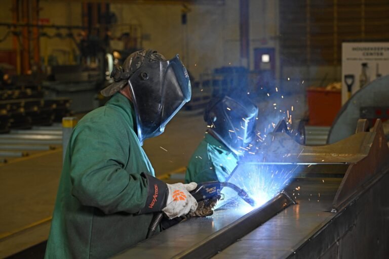 Manitowoc Expands Rapid Response Shop and Strengthens Aftermarket Support Welder in a green protective jacket and helmet welding metal, sparks flying in an industrial workshop.