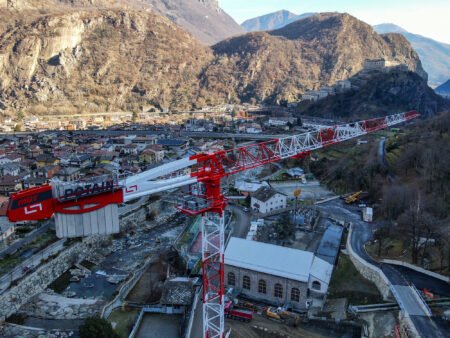 Large red construction crane looms over a valley town with a fortress on a hill and distant mountains in the background.”,