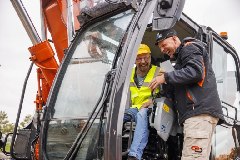 Two construction workers in a cab of an orange excavator, wearing safety gear and sharing a notebook while smiling at each other