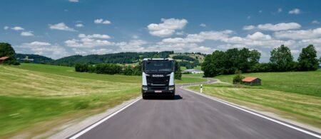 Scania-electrification-France-plantjpg - Machines Wiki Black Scania truck driving toward the viewer on a rural two-lane road, green fields and blue sky around it on a sunny day.