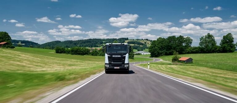 Black Scania truck driving toward the viewer on a rural two-lane road, green fields and blue sky around it on a sunny day.