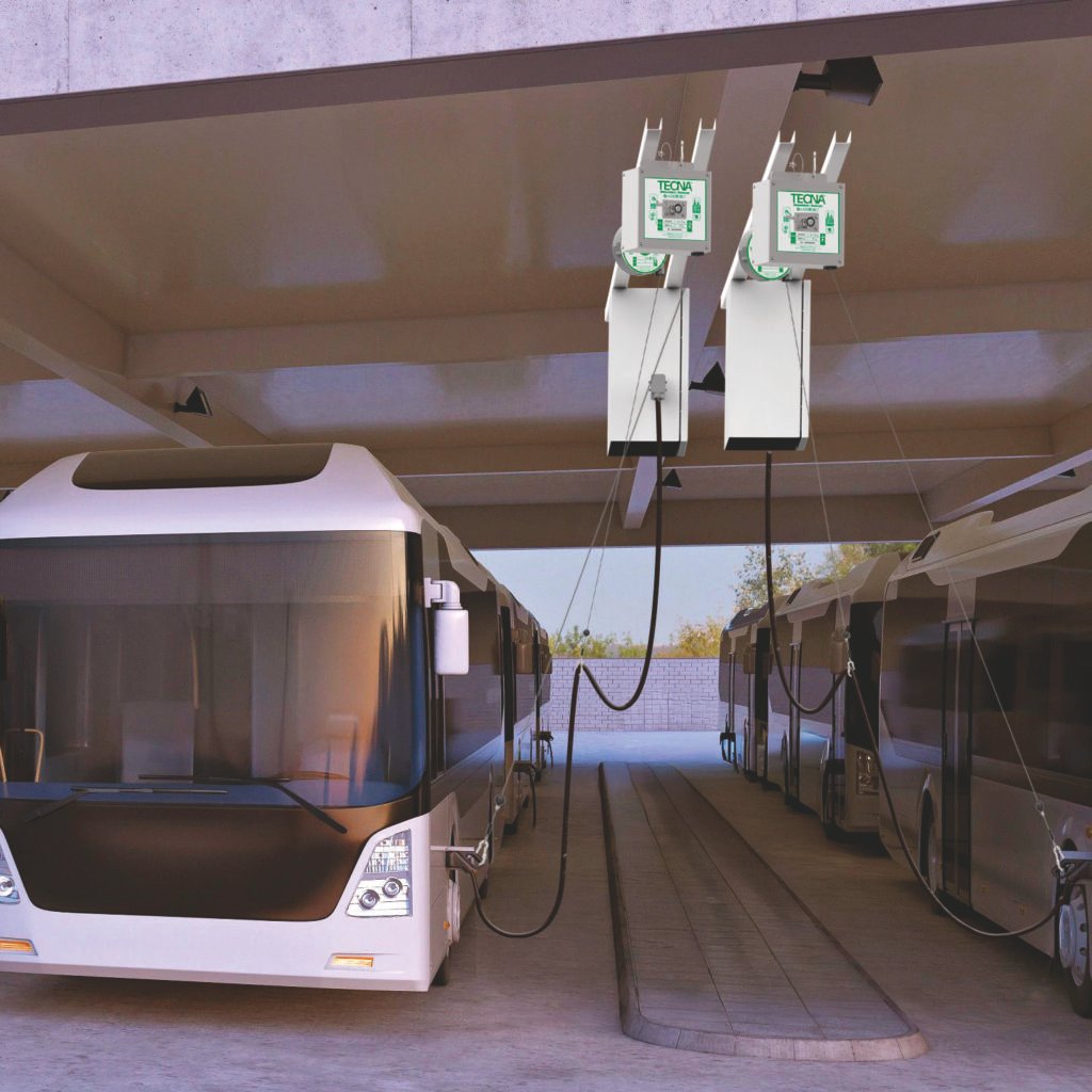 Two white buses are parked under a concrete overhang, connected to overhead electric charging units labeled TECNA with green screens above each vehicle objective: EV charging.