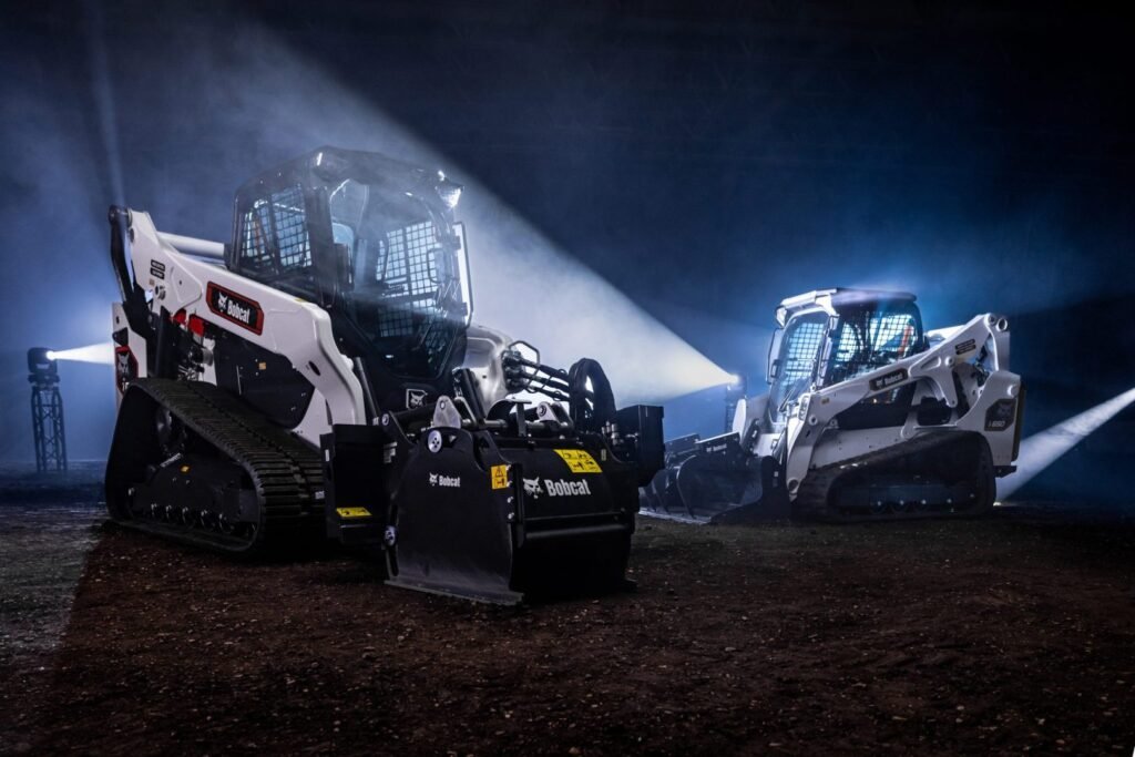Classic or Pro? Bobcat simplifies compact loader selection Two Bobcat skid steer loaders with tracks sit on a dirt floor under dramatic blue stage lighting in a dark indoor space.