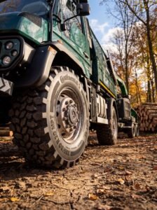 Close-up of a heavy-duty green logging truck tire on a muddy dirt road with stacked logs and autumn trees in the background.