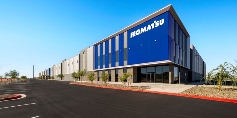 Komatsu to Open New Parts Distribution Facility in Arizona Modern Komatsu distribution center with a blue-panel facade and glass entrance under a clear blue sky