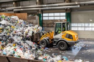 Front-end loader (Liebherr) piles through a large indoor garbage refuse pile next to a warehouse door and waste bins, showing an active recycling/dumping site interior.