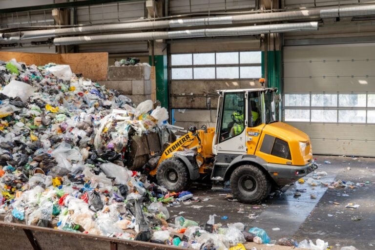 liebherr-L507-E-G8_300dpi-scaledjpg - Machines Wiki Front-end loader (Liebherr) piles through a large indoor garbage refuse pile next to a warehouse door and waste bins, showing an active recycling/dumping site interior.