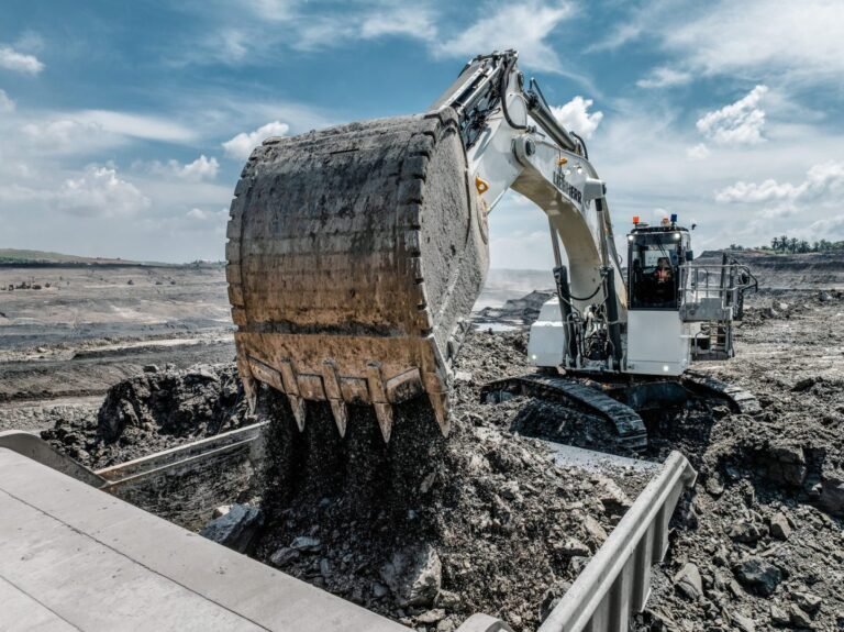 Large excavator dumps muddy earth into a trench at a barren mining site under a blue sky. Best for an informative image showing earthmoving work in a quarry/field site.