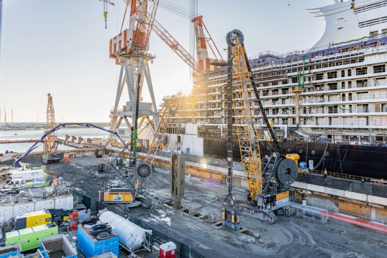 liebherr-rcm_2_300dpi-scaledjpg - Machines Wiki Crane-equipped shipyard at the water with a partially built cruise liner and heavy construction equipment in the foreground at sunset sun glare in the background