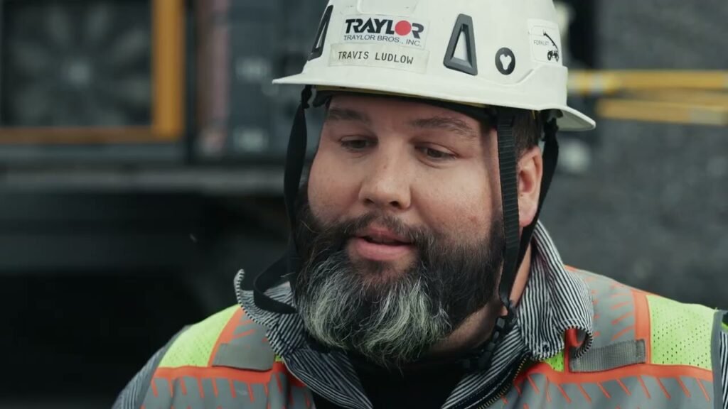 Construction worker wearing a white safety helmet with Taylor label and a neon-orange reflective vest, looking downward.