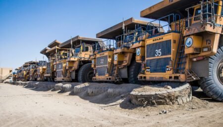 Row of large yellow mining dump trucks parked on a dusty construction site under a clear blue sky.