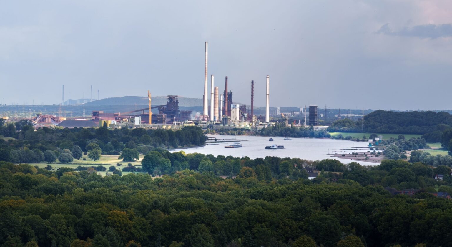 Industrial plant with tall smokestacks along a river, seen behind a lush, tree-filled foreground and small boats on the water