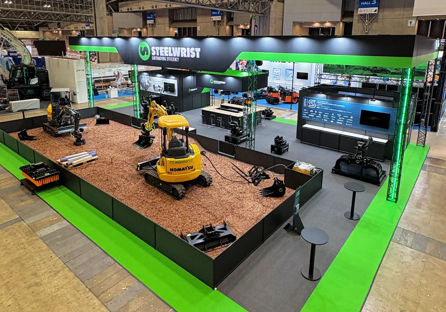 1-Steelwrist-biggest-stand-ever-at-CSPI-EXPO_2026-scaledwebp - Machines Wiki Wide exhibition booth for Steelwrist with green and black branding, showing excavators and display screens inside a trade show hall