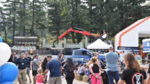 1783023752_63948_en_0196b_66097_falcon-equipment-crane-rodeopng - Machines Wiki Crowd at an outdoor trade show with a red boom crane, blue truck, tents, and pine trees in the background.