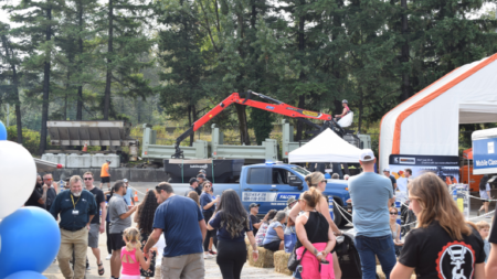 1783023752_63948_en_0196b_66097_falcon-equipment-crane-rodeopng - Machines Wiki Crowd at an outdoor trade show with a red boom crane, blue truck, tents, and pine trees in the background.