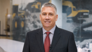 1783097883_63957_en_b2e2a_66106_mike-ross-hd-hyundai-presidentpng - Machines Wiki Smiling man in a dark pinstripe suit and red tie, with a lapel microphone, in a bright office with construction machinery imagery on a glass wall.