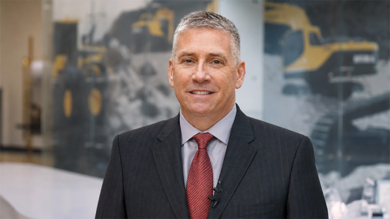 Smiling man in a dark pinstripe suit and red tie, with a lapel microphone, in a bright office with construction machinery imagery on a glass wall.