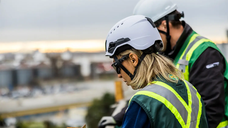 1783101561_63954_en_b6e81_66103_ccsc-type2-hard-hatpng - Machines Wiki Two construction workers in white hard hats and high-visibility vests look down at a work area on a blurred construction site.