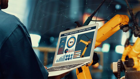 1783105208_63959_en_7b8e6_66109_cat-workforce-initativepng - Machines Wiki Person in a blue shirt holds a laptop displaying a smart industrial control panel with charts while a yellow robotic arm operates in the background, in a factory setting.