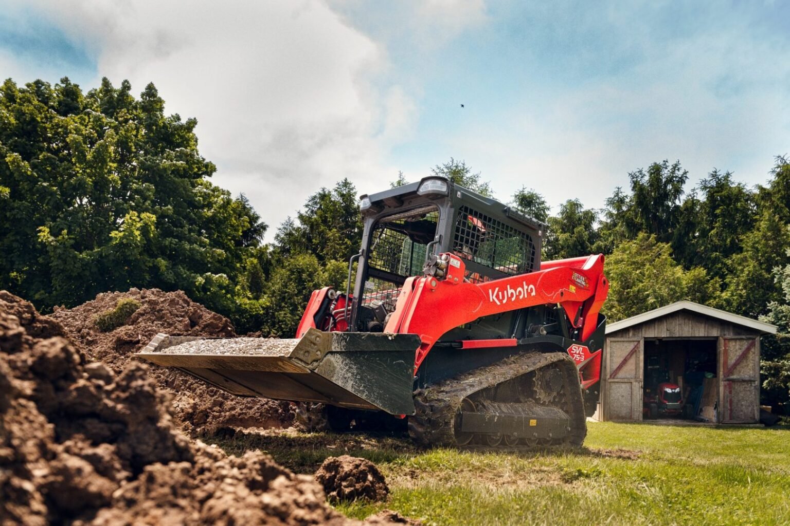 Red Kubota skid-steer loader with bucket raised, moving soil on a grassy construction site near a shed and trees in the background.
