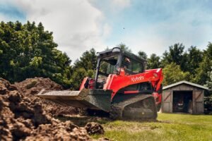 1783397246_HERO-IMAGE_240529-RV-UD-KBT_-6-scaledjpg - Machines Wiki Red Kubota skid-steer loader with bucket raised, moving soil on a grassy construction site near a shed and trees in the background.