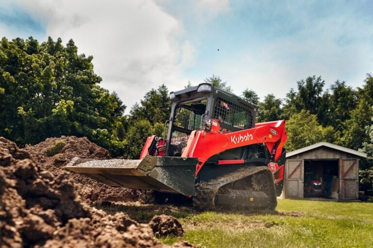 1783397246_HERO-IMAGE_240529-RV-UD-KBT_-6-scaledjpg - Machines Wiki Red Kubota skid-steer loader with bucket raised, moving soil on a grassy construction site near a shed and trees in the background.