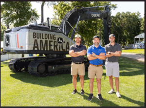 1783435322_John-Deere-Building-America-Contestpng - Machines Wiki Three men stand with a large excavator labeled 'BUILDING AMERICA' at an outdoor park event on a sunny day.