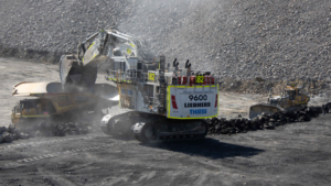 1783616784_63978_en_2ca88_66136_thiess-liebherr-r9600-mining-excavatorpng - Machines Wiki Large excavator loads coal into a dump truck at an open-pit mine, with a rocky wall and dust in the air.
