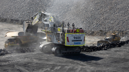 1783616784_63978_en_2ca88_66136_thiess-liebherr-r9600-mining-excavatorpng - Machines Wiki Large excavator loads coal into a dump truck at an open-pit mine, with a rocky wall and dust in the air.