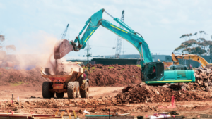 1783628005_63977_en_0f803_66135_unsplash-construction-fleetpng - Machines Wiki Teal excavator loads dirt into a yellow dumping truck at a construction site, dust rising as earth is moved.