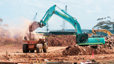 1783628005_63977_en_0f803_66135_unsplash-construction-fleetpng - Machines Wiki Teal excavator loads dirt into a yellow dumping truck at a construction site, dust rising as earth is moved.