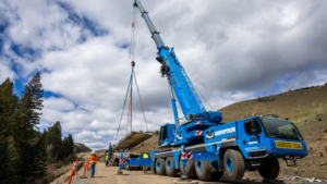 Blue heavy-lift crane on a dirt road lowers a large concrete slab while workers guide it during road construction in a mountainous area under cloudy skies.