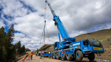 Blue heavy-lift crane on a dirt road lowers a large concrete slab while workers guide it during road construction in a mountainous area under cloudy skies.