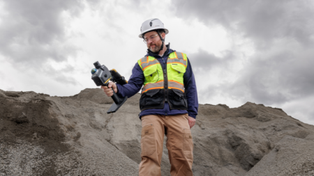 1784140017_64001_en_dea84_66166_topcon-gvipng - Machines Wiki Surveyor wearing a hard hat and high‑visibility vest using a handheld laser/scanner on a dirt pile outdoors